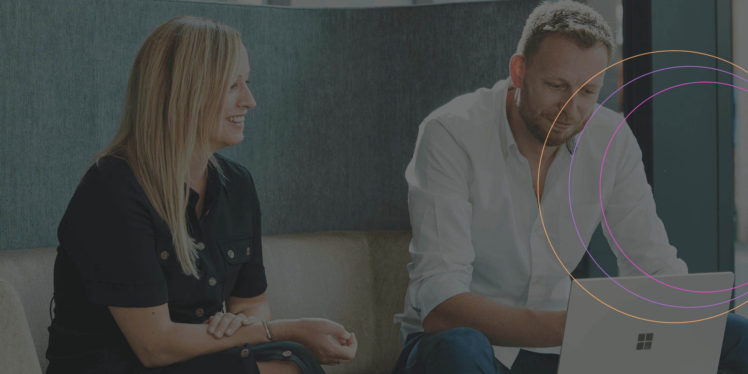 Smiling man and woman having a discussion in an office meeting on sofa with grey walls and a slightly open door in the background and a laptop in front of them