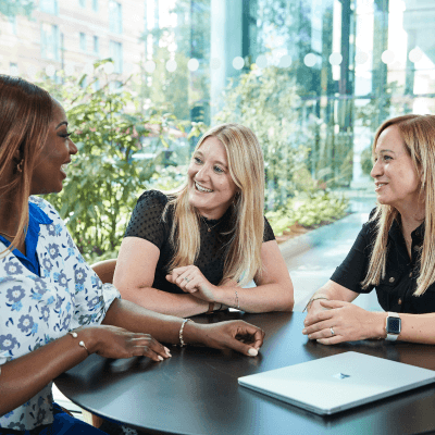 Three smiling women in an office sitting at a table having a meeting with wide windows in the background and a laptop on the table