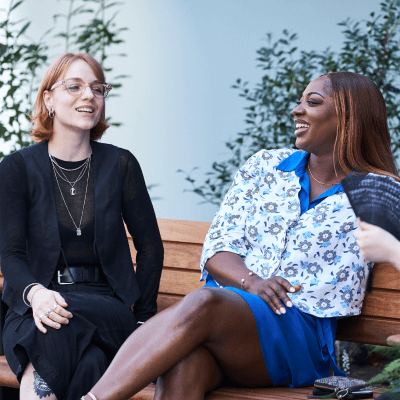 Two women sitting on a bench laughing and smiling and having a discussion with green plants in the background