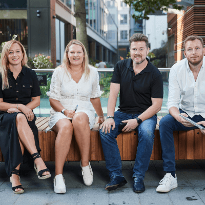 Two men and two women smiling and sitting on a bench in the sunshine with office blocks in the background