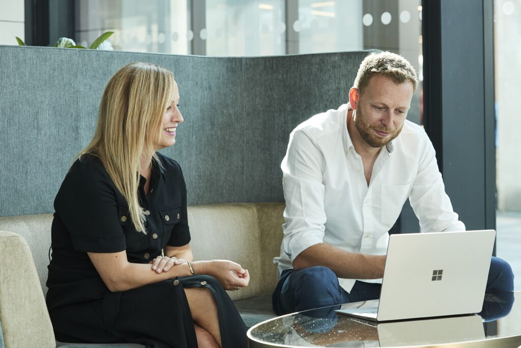 Smiling man and woman having a discussion in an office meeting on sofa with grey walls and a slightly open door in the background and a laptop in front of them